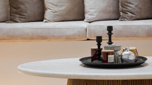 Ein modernes Wohnzimmer mit einem runden Holztisch, einem weißen Sofa mit beige farbenen Kissen und einer kleinen Ablage mit einer Kerze, einer Flasche und einem Metallkrug.