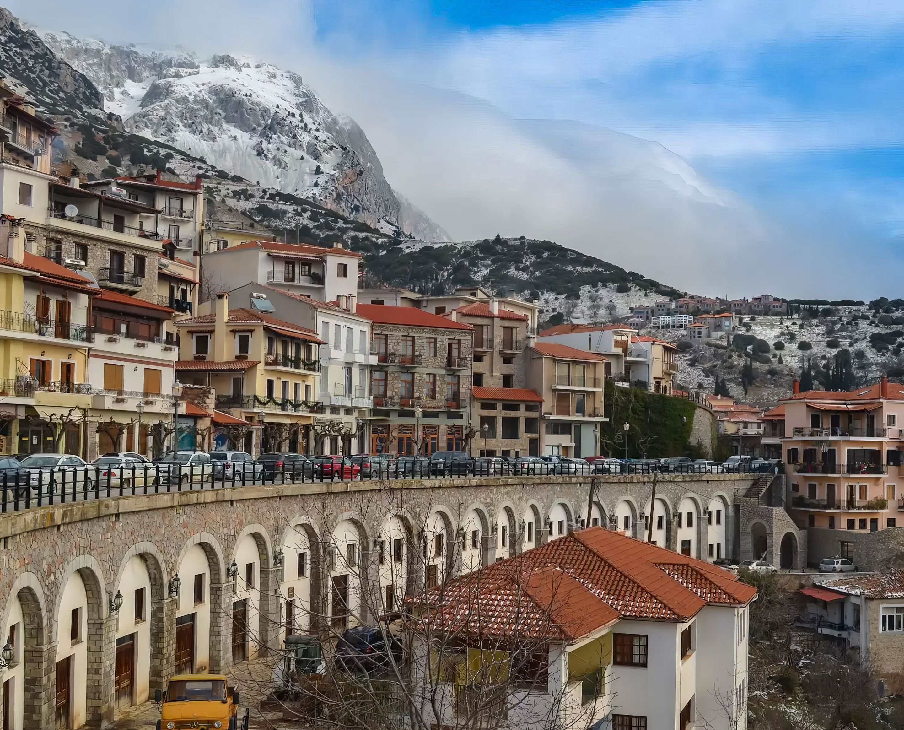 A town with a prominent stone bridge, buildings with red-tiled roofs, and a mountainous backdrop.