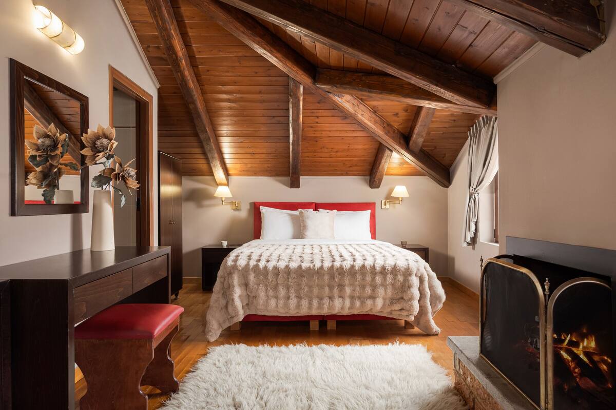 Une chambre avec un plafond en bois, un lit avec une tête de lit rouge, une cheminée et un miroir décoré de motifs floraux.