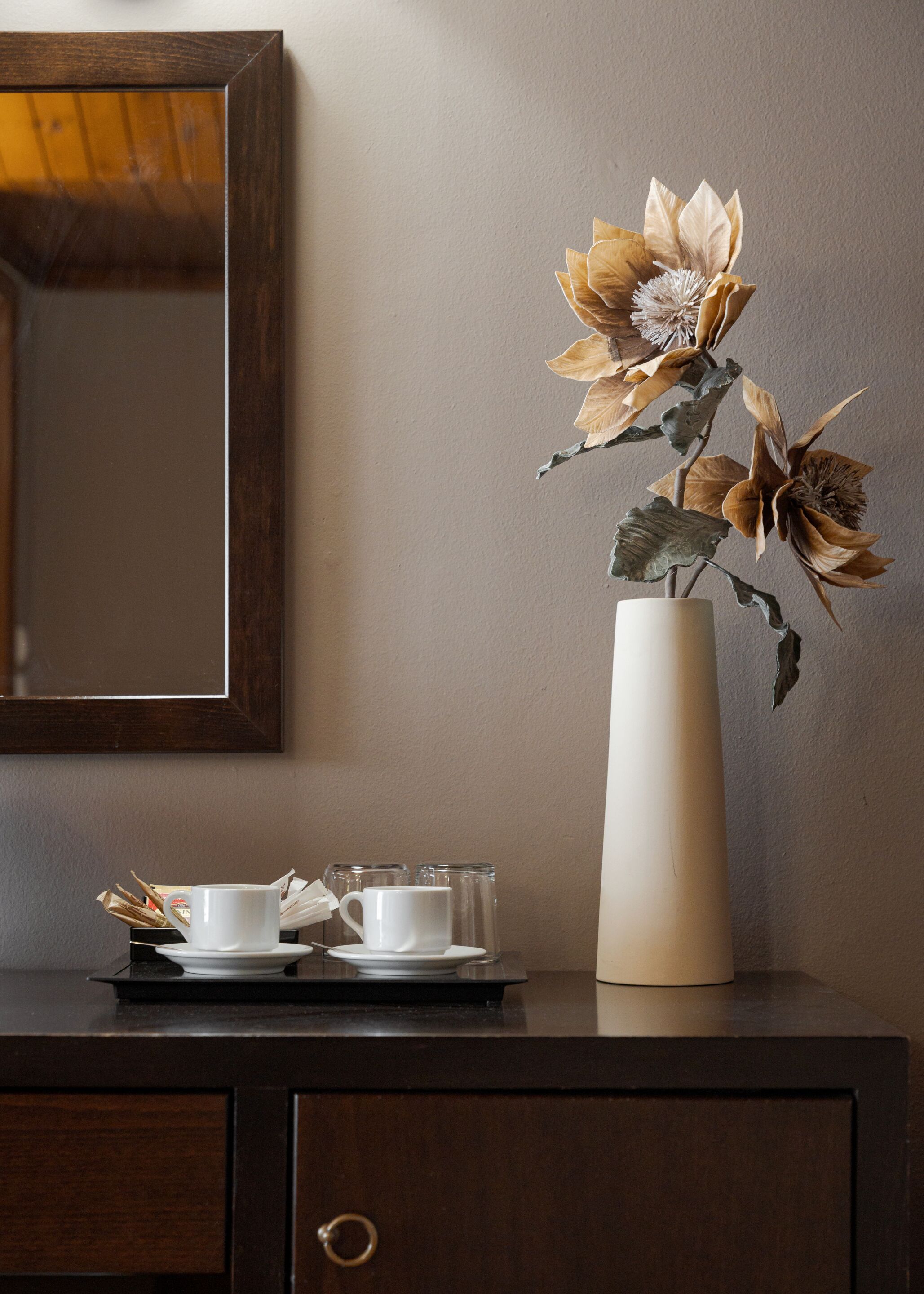 A vase with dried flowers on a wooden table next to a mirror and a tray with cups and saucers.