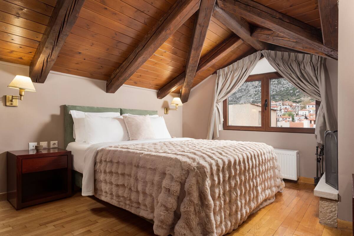 Une chambre avec un plafond en bois, un lit recouvert d’une couverture moelleuse, une table de chevet, une fenêtre avec des rideaux et une vue sur les bâtiments à l’extérieur.