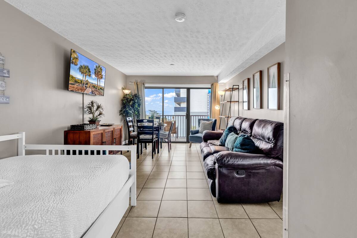 A modern living room with a bed, a leather sofa, a dining table with chairs, and a TV mounted on the wall.