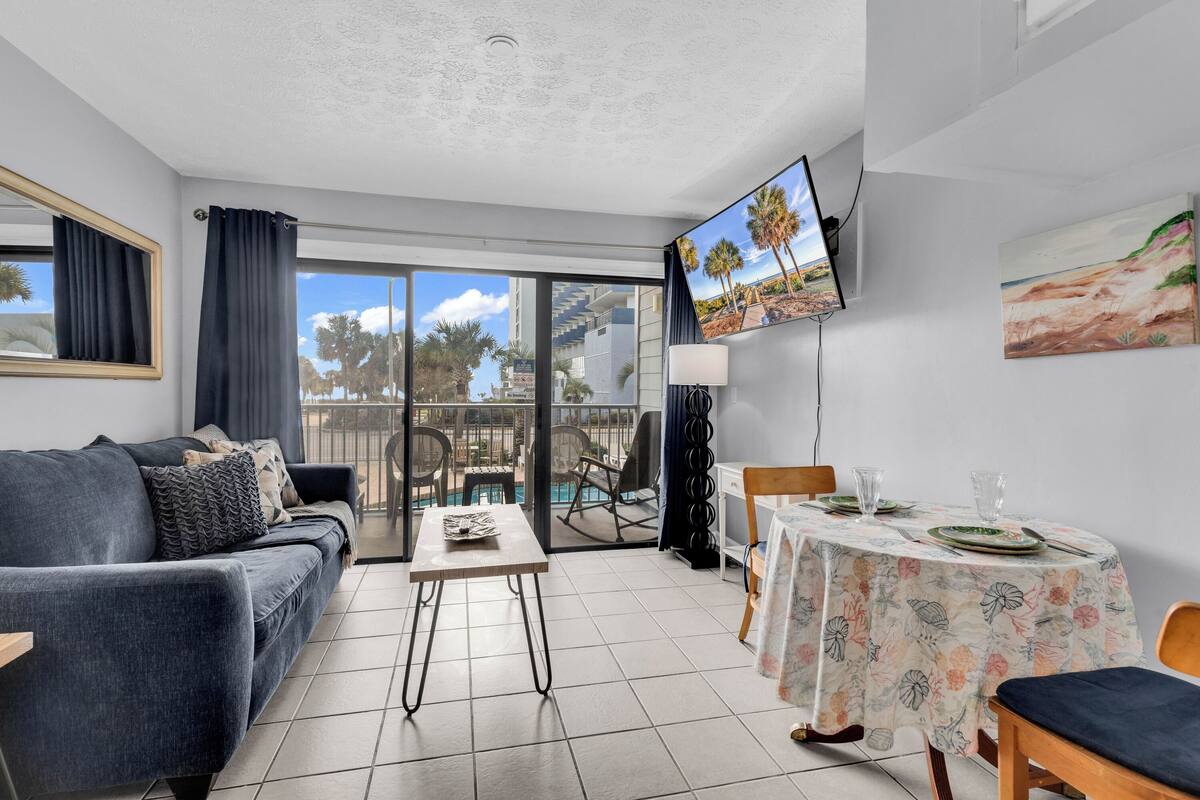 A modern living room with a sofa, a dining table set for two, a TV mounted on the wall, and a balcony with a pool view.