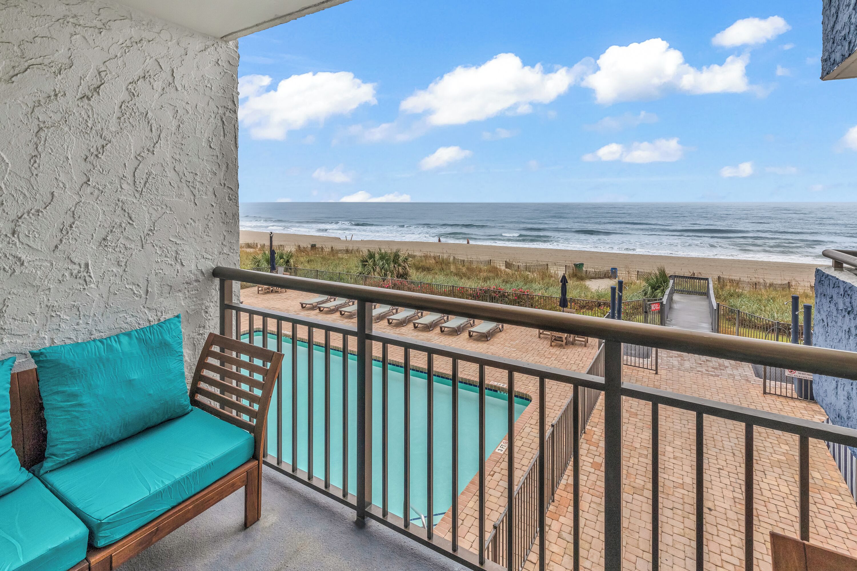 A balcony with a view of the beach and ocean, a pool, and a wooden bench with teal cushions.