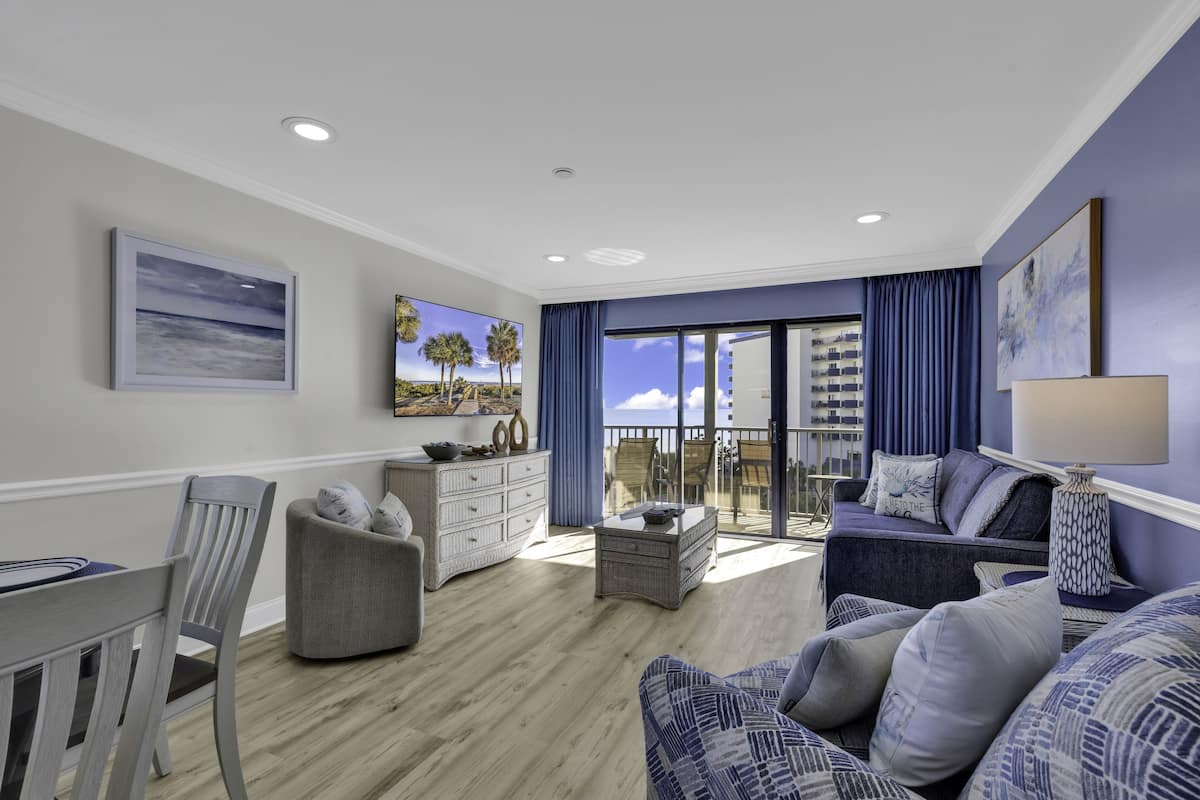 A spacious living room with a balcony, a dining area, and a view of the ocean.