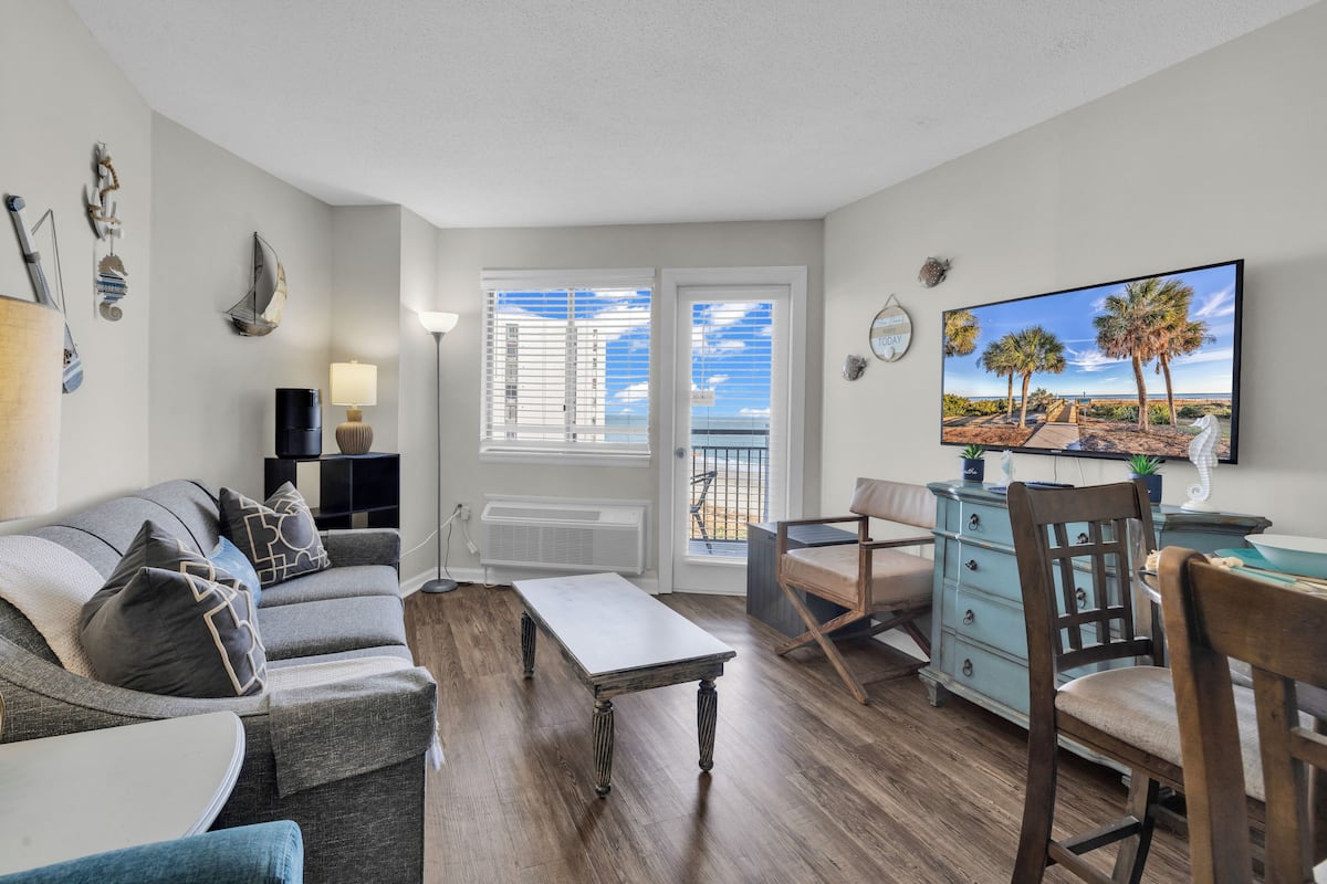 A living room with a sofa, a coffee table, a TV, and a view of the outdoors through a door.