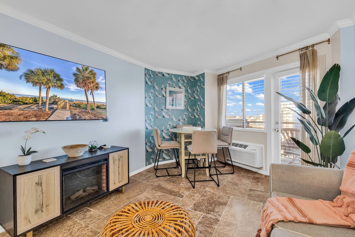 A living room with a fireplace, a dining area, and a large window with a view of palm trees.
