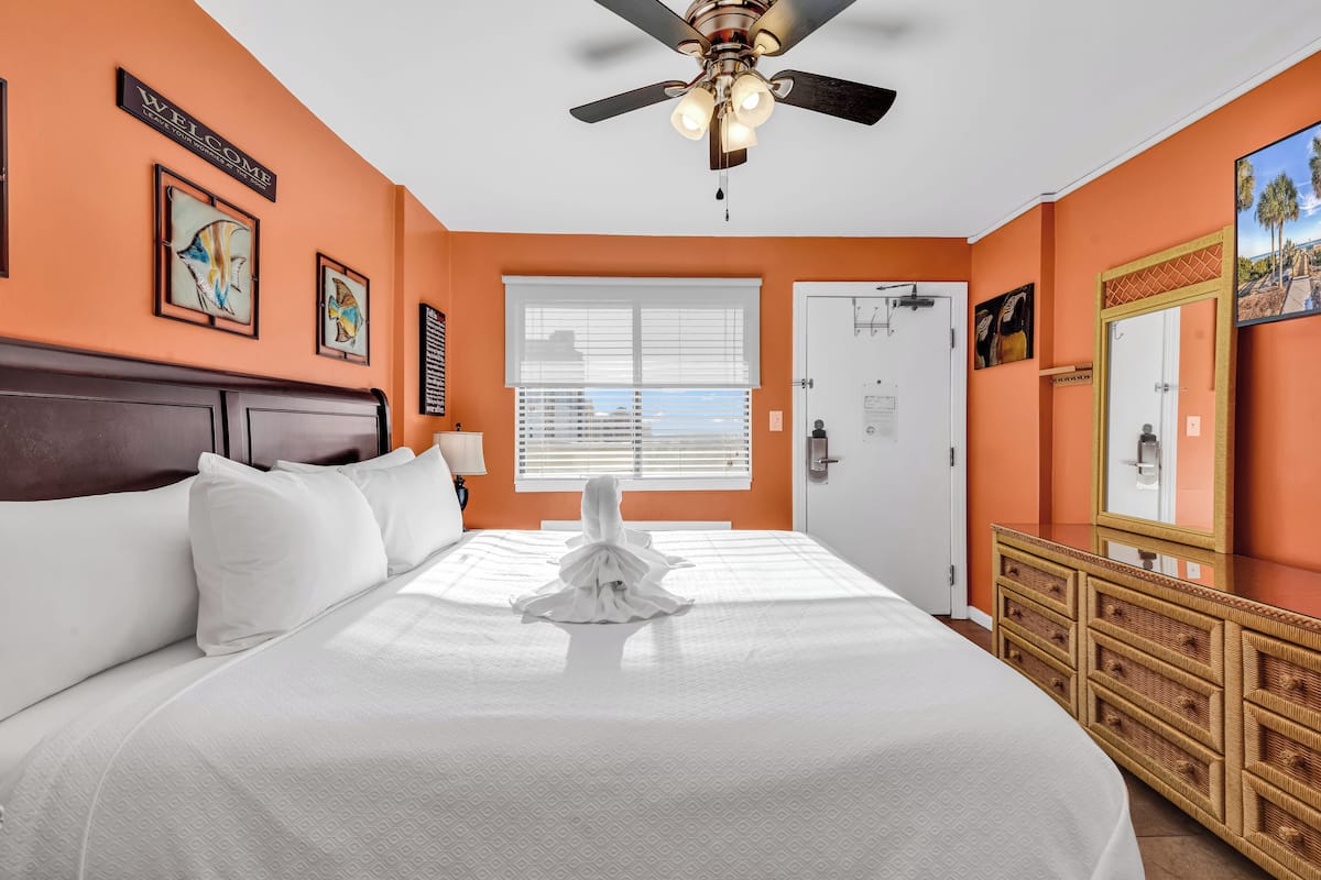 A hotel room with a large bed, a ceiling fan, a window with blinds, a wooden dresser, and artwork on the walls.