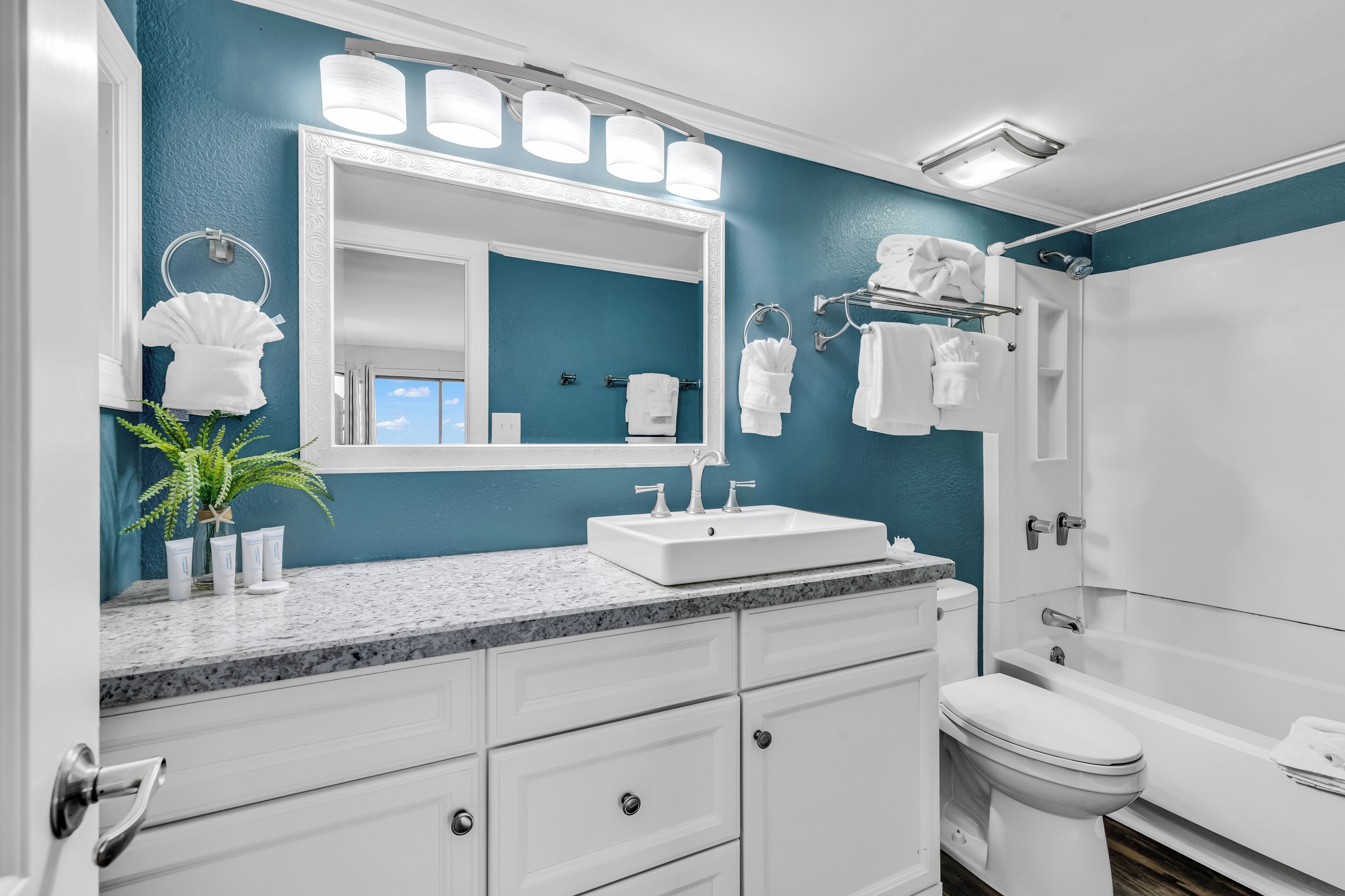 A bathroom with a large mirror, white sink, and a toilet. There are towels hanging on a rack and a plant on the counter.