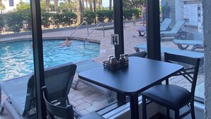 A poolside area with a table and chairs, a pool, and a view of palm trees and a building in the background.