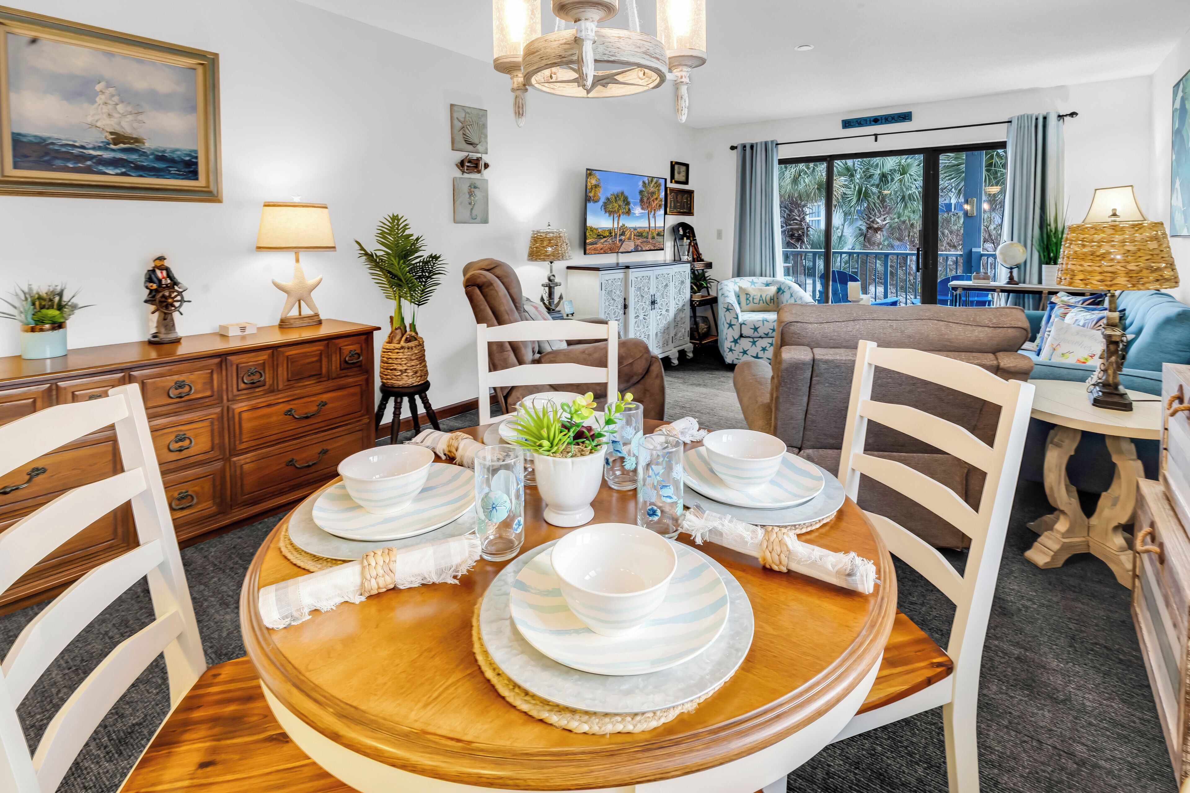 A dining area with a wooden table set for four, surrounded by white chairs. There is a large window with a view of palm trees and a balcony.