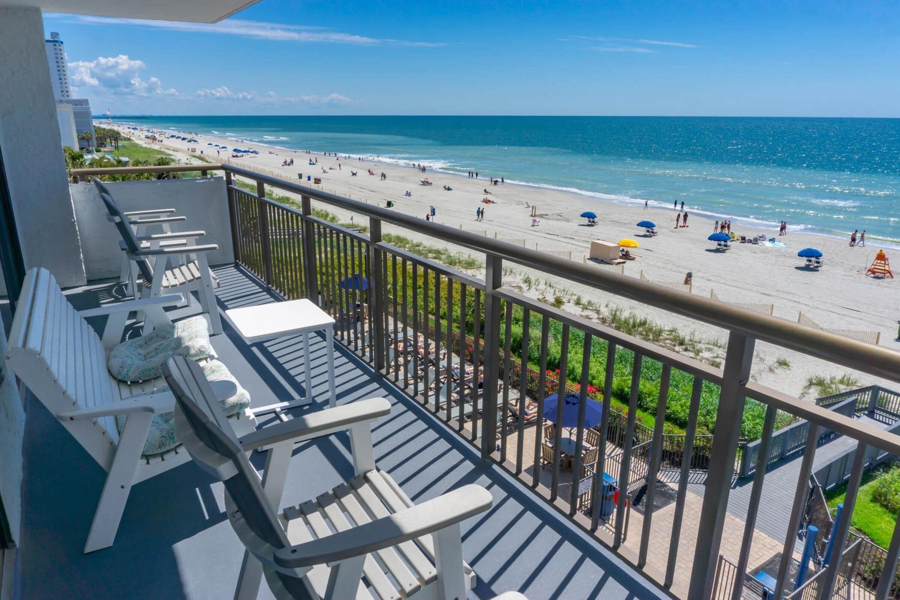 A balcony with white Adirondack chairs and a view of a beach with umbrellas and people.