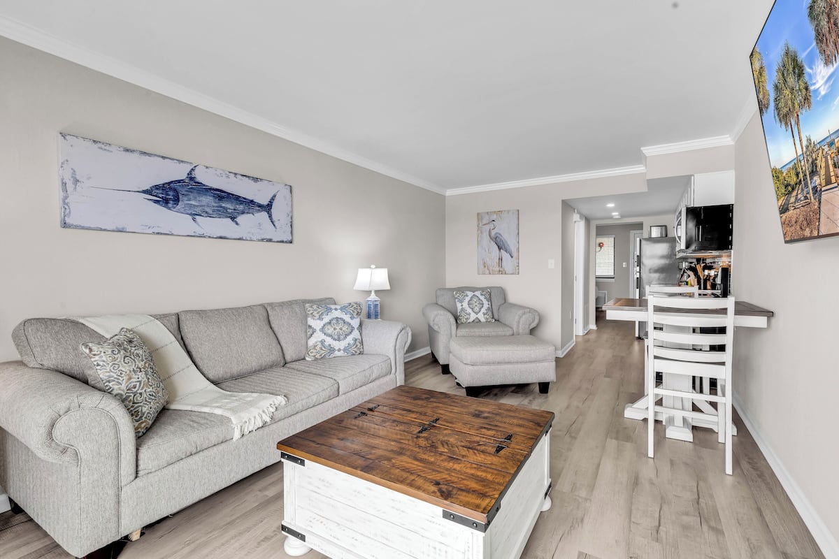 A living room with a grey sofa, a coffee table, and a chair. There is a painting of a bird on the wall.
