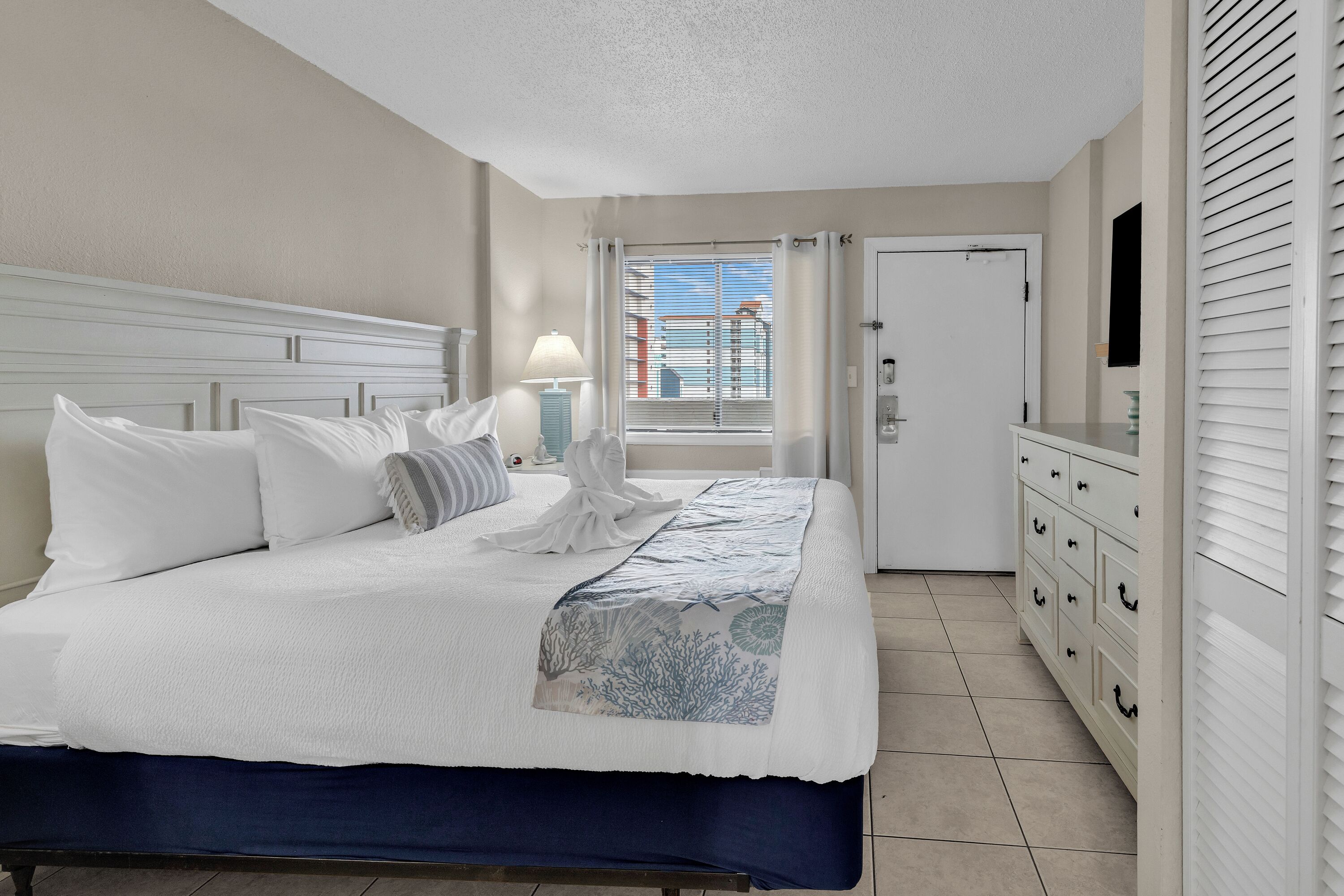 Une chambre d’hôtel avec un lit, une commode, une télévision et une fenêtre avec des stores.