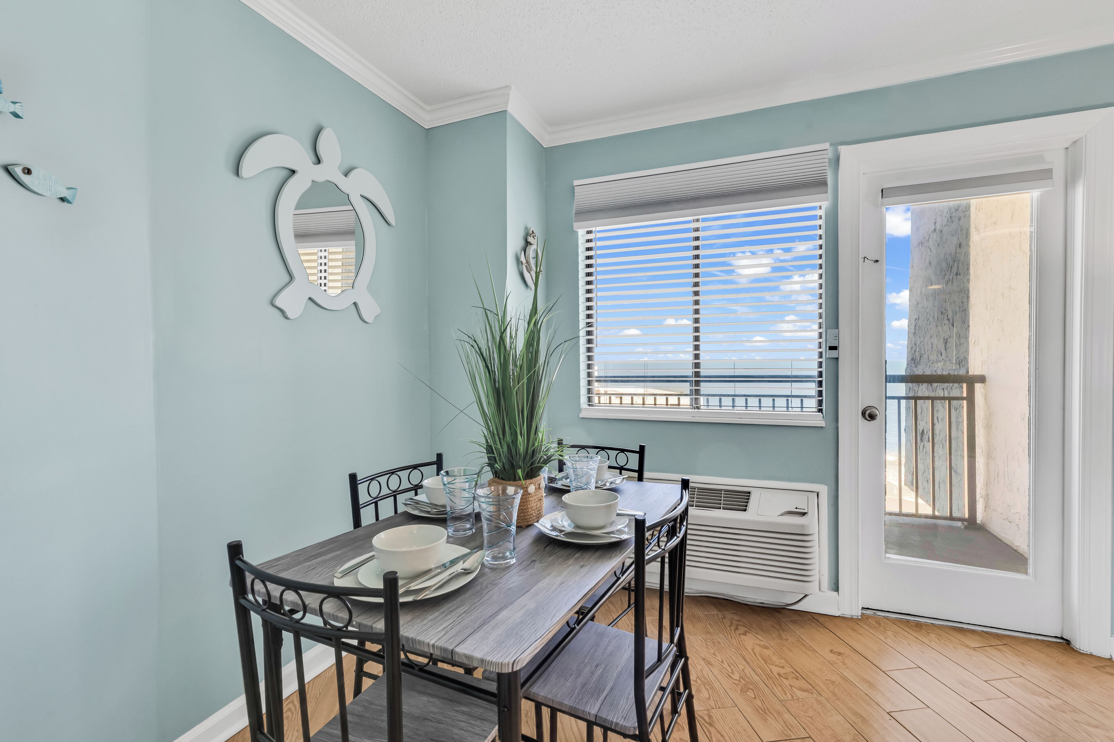 A dining area with a table set for four, a potted plant, and a window with blinds.