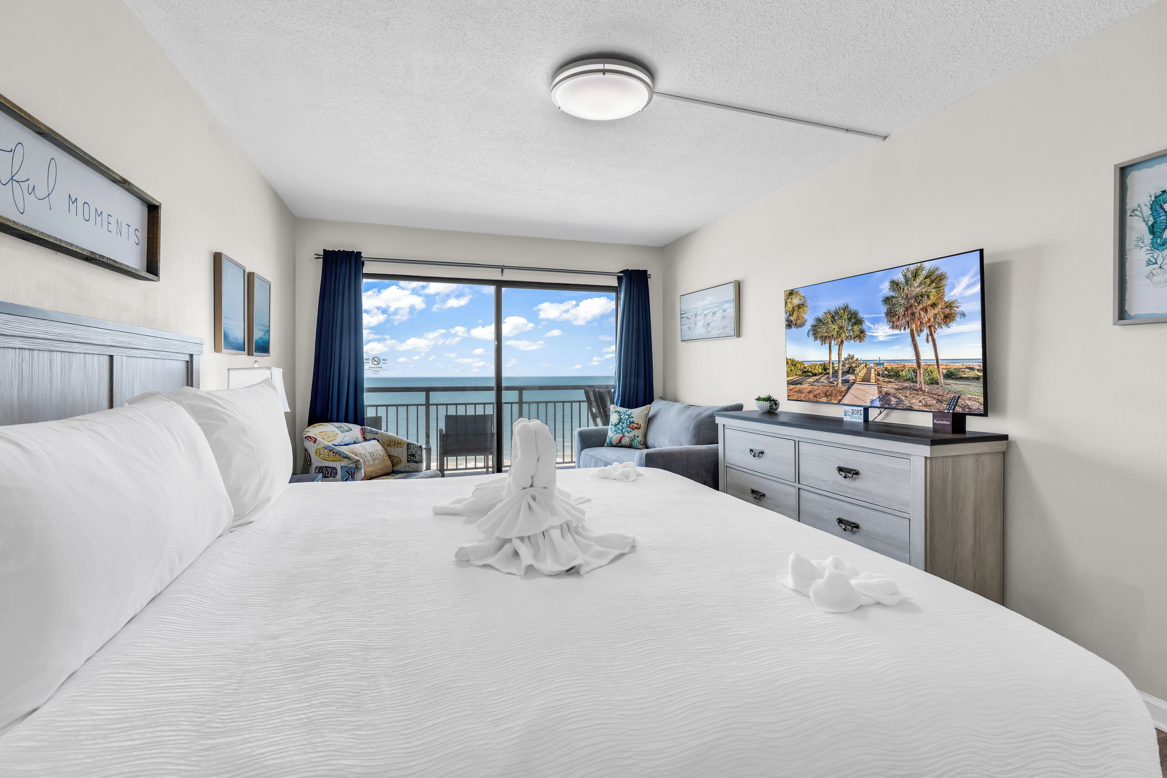 A hotel room with a large bed, a TV, a balcony with a view of the ocean, and a framed picture on the wall.