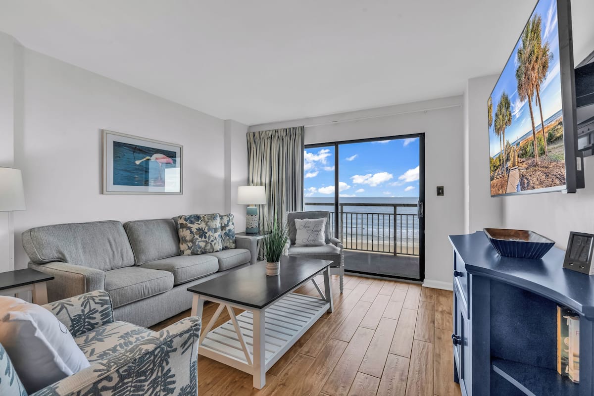 A modern living room with a grey sofa, a coffee table, and a view of the ocean through large windows.