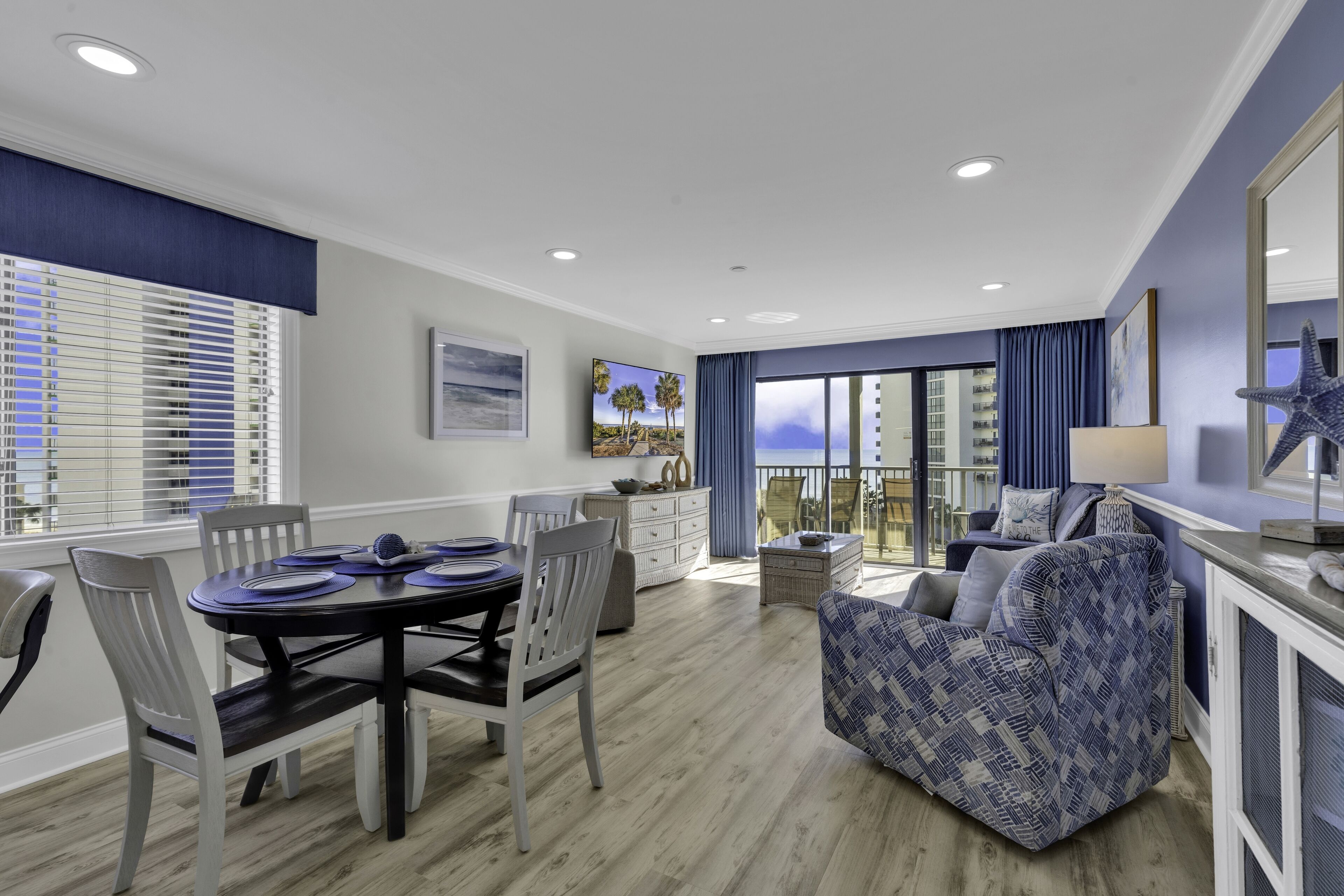 A modern living room with a dining area, featuring a round table, chairs, a sofa, and a large window with blinds.