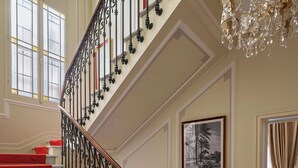 Una imponente escalera con peldaños alfombrados de rojo y barandillas de hierro forjado negro, que conduce a una sala con candelabro y un cuadro en la pared.