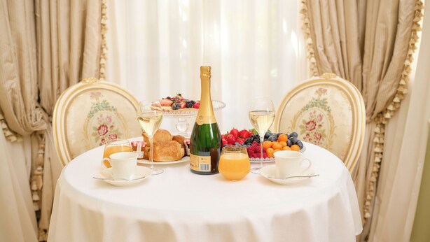 Una mesa servida con champán, fruta, pasteles y tazas, rodeada de sillas ornamentadas en una habitación con cortinas.