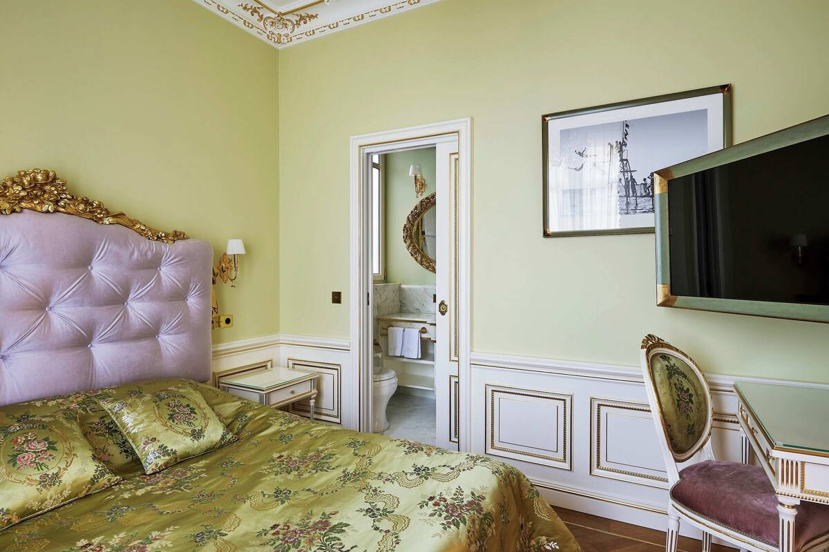 Une chambre à coucher avec une tête de lit capitonnée, un lit recouvert d’une literie à motifs floraux, une chaise avec un siège rembourré, un tableau encadré accroché au mur et un miroir placé dans l’embrasure de la porte.