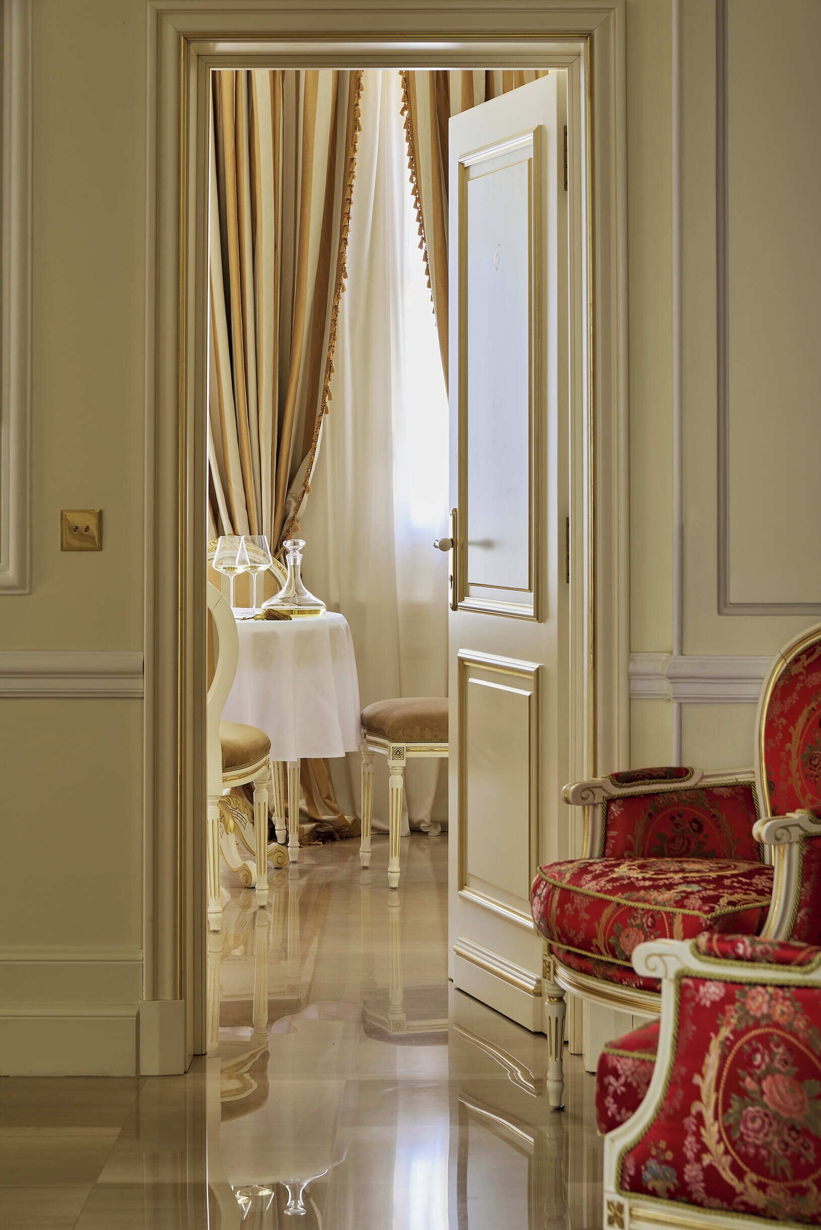 Una habitación con piso pulido, una mesa con mantel blanco y dos sillas con tapicería roja.