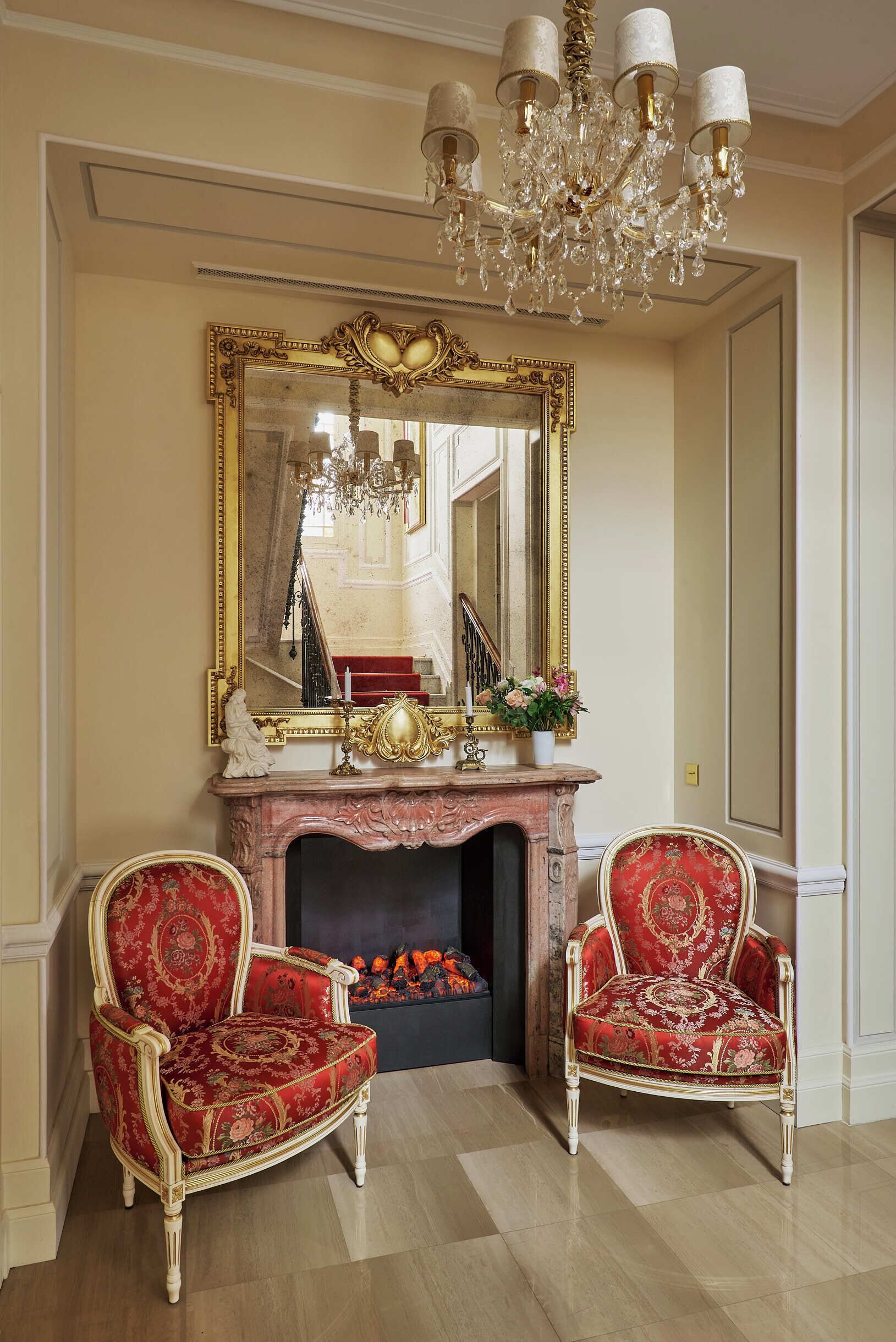 A room with a grand mirror, two ornate chairs, a fireplace, and a chandelier.