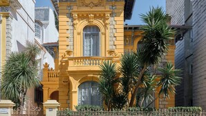 Un edificio amarillo con detalles arquitectónicos ornamentales, que incluyen un balcón y ventanas amplias.