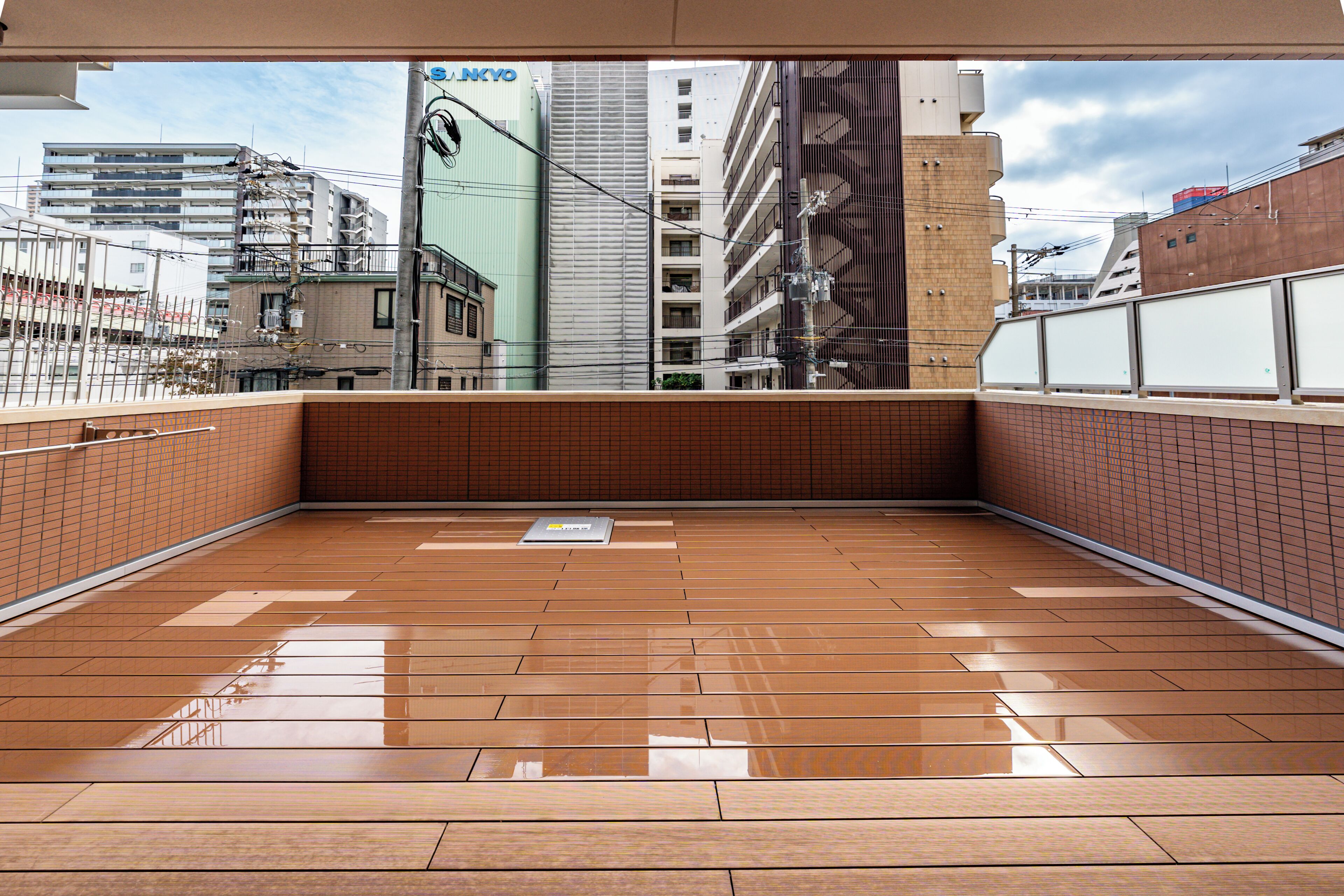 A spacious balcony with a tiled floor, overlooking a cityscape with various buildings and a clear sky.