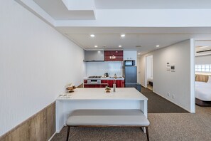 Un estrecho pasillo de hotel con una kitchenette, una pequeña mesa de comedor y una cama en la habitación contigua.