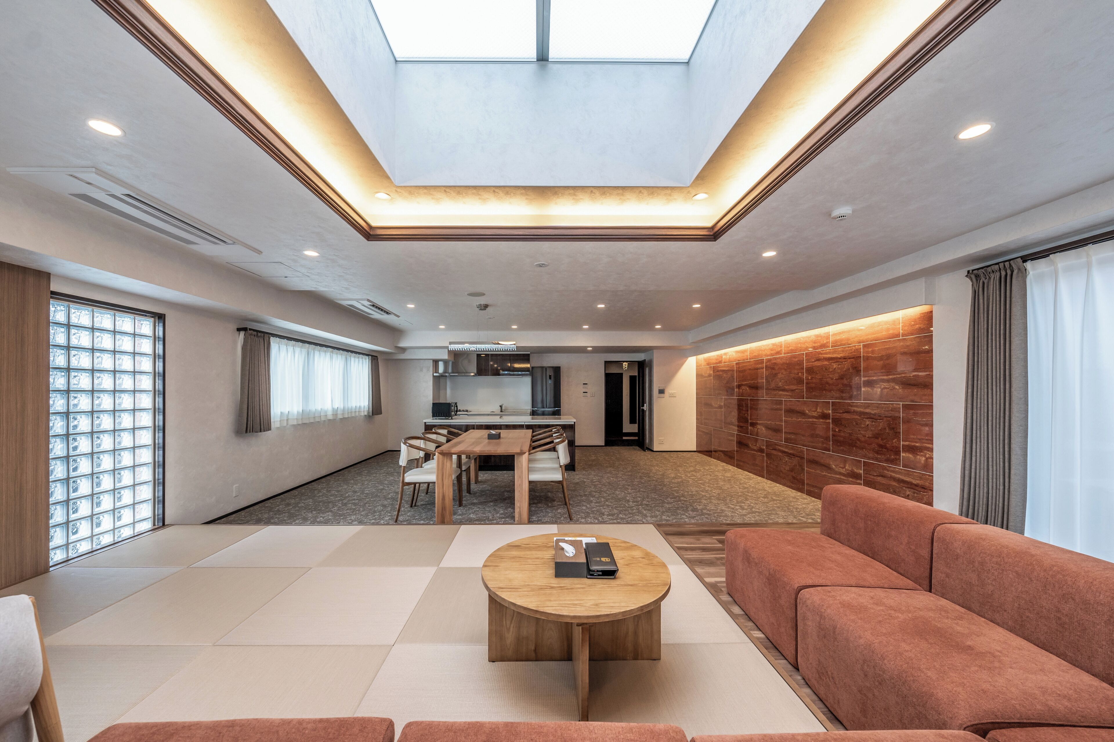 A spacious living area with a high ceiling, a central wooden coffee table, a sofa, and a dining area with a kitchen in the background.