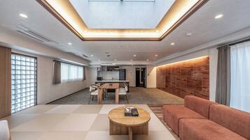 A spacious living area with a high ceiling, a central wooden coffee table, a sofa, and a dining area with a kitchen in the background.