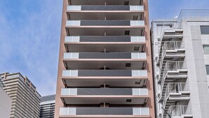 A multi-story building with a modern architectural design, featuring balconies on each floor, a ground-level entrance, and a parking area visible in the foreground. - Apartment Hotel 11 Namba Motomachi (Osaka)