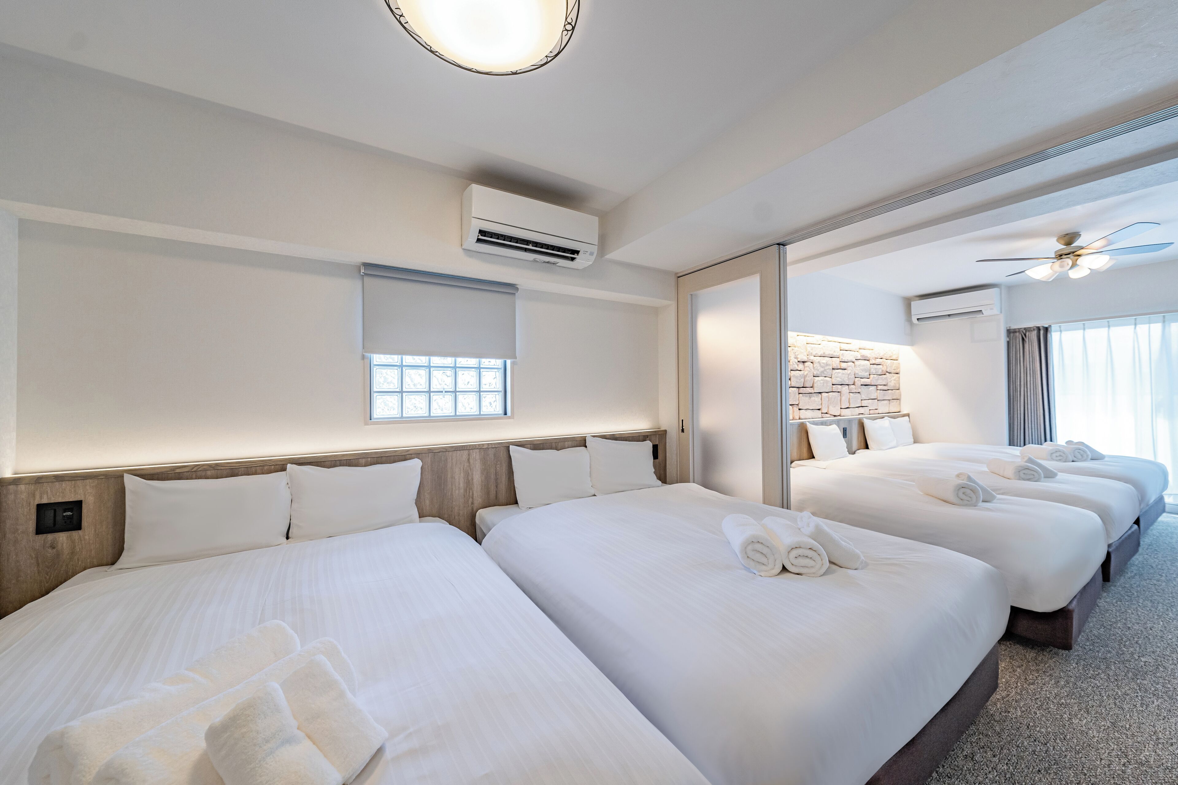 A hotel room with three beds, an air conditioning unit, a window with blinds, and a ceiling fan.