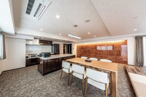 Eine moderne Küche mit einer zentralen Kochinsel, ein Essbereich mit einem Holztisch und weißen Stühlen sowie ein Wohnbereich mit großem Fenster und einer Ziegelwand.