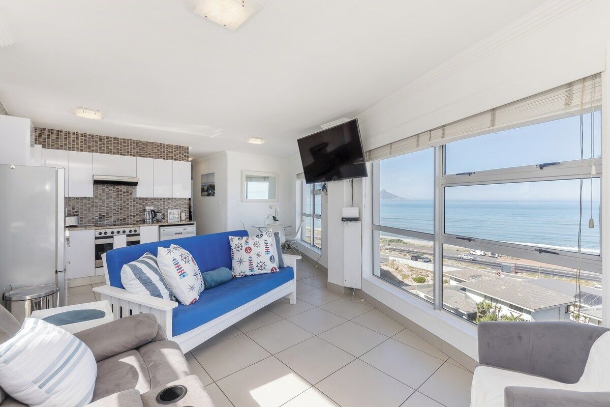 A modern living room with a blue sofa, a flat-screen TV mounted on the wall, and a large window offering a view of the sea.