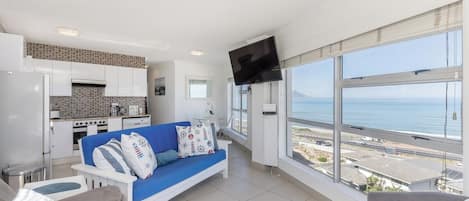 Un salon moderne avec un canapé bleu, un téléviseur à écran plat fixé au mur et une grande fenêtre donnant sur la mer.