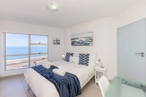 A bedroom with a large window, a bed with a blue blanket, a bedside table with a lamp, and a painting on the wall.