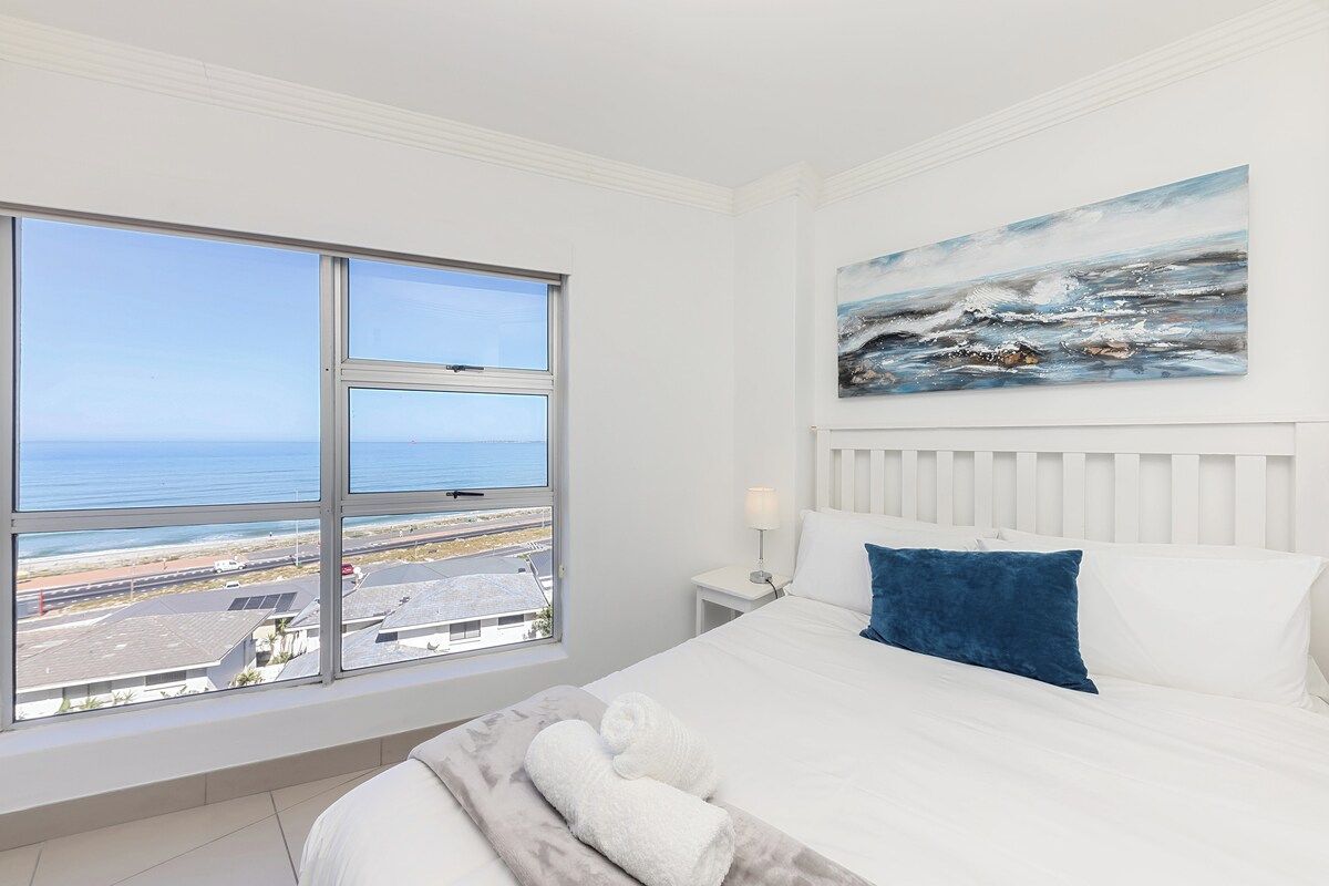 A hotel room with a bed, a painting, a nightstand, and a window with a view of the sea and buildings.