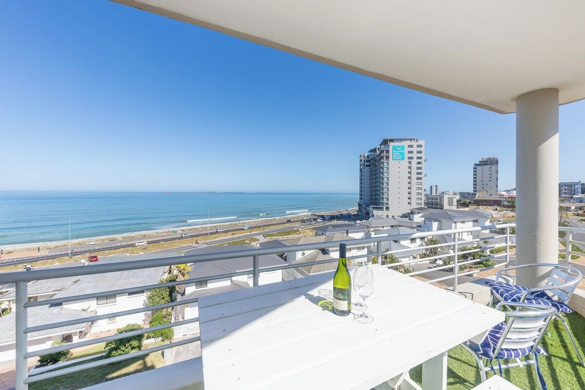 A balcony with a view of the beach and cityscape, a table with a bottle and glass, and a chair with a blue cushion.