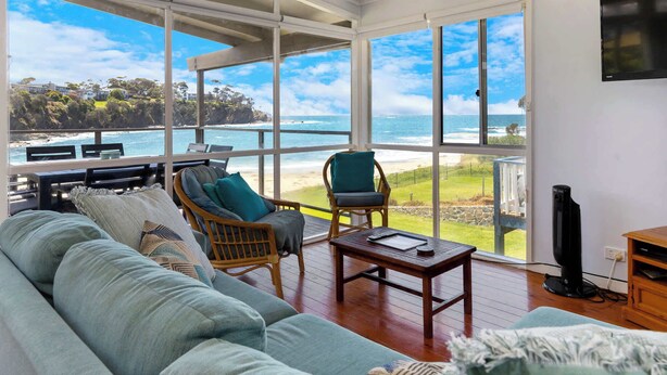 Ein geräumiges Wohnzimmer mit großem Fenster mit Blick auf Strand und Meer. Es gibt ein Sofa, zwei Sessel und einen Couchtisch.