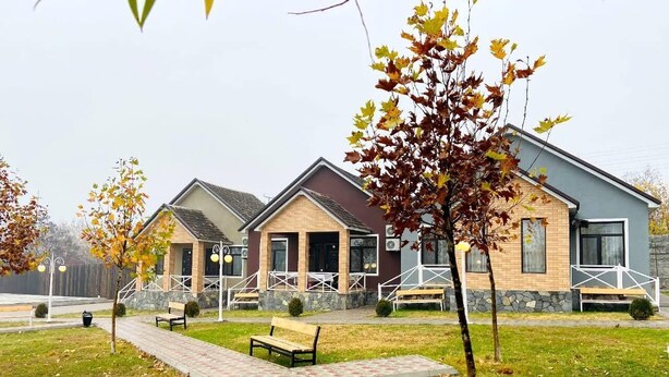 A row of modern buildings with a paved walkway and benches, surrounded by grass and trees with autumn leaves. - Tulpan Hotel (Sheki)
