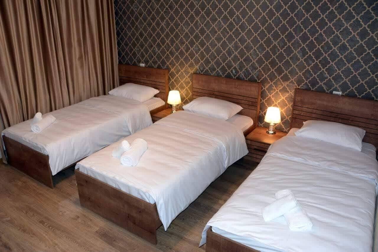 Et hotelværelse med tre enkeltsenge, hver med en træsenggavl og en sengebordlampe.