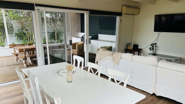 A modern living room with a white table and chairs, a sofa, and a TV mounted on the wall. - Saltwater Bungalow - WiFi (Robe)