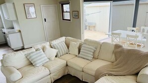 A living room with a beige sectional sofa, a small kitchen area, and a sliding glass door leading to an outdoor patio. - Saltwater Bungalow - WiFi (Robe)