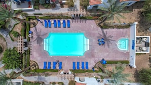 Vista aérea de un hotel con piscina central rodeada de sillas reclinables azules, una piscina más pequeña y palmeras.