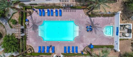 Vista aérea de un hotel con piscina central rodeada de sillas reclinables azules, una piscina más pequeña y palmeras.