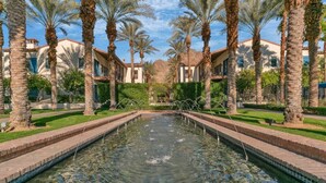 A rectangular pool with clear water, surrounded by palm trees and buildings with red-tiled roofs. - Overlook Oasis C | Studio Legacy Villa (La Quinta)