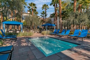 A swimming pool with blue lounge chairs and umbrellas, surrounded by palm trees and greenery. - Overlook Oasis C | Studio Legacy Villa (La Quinta)