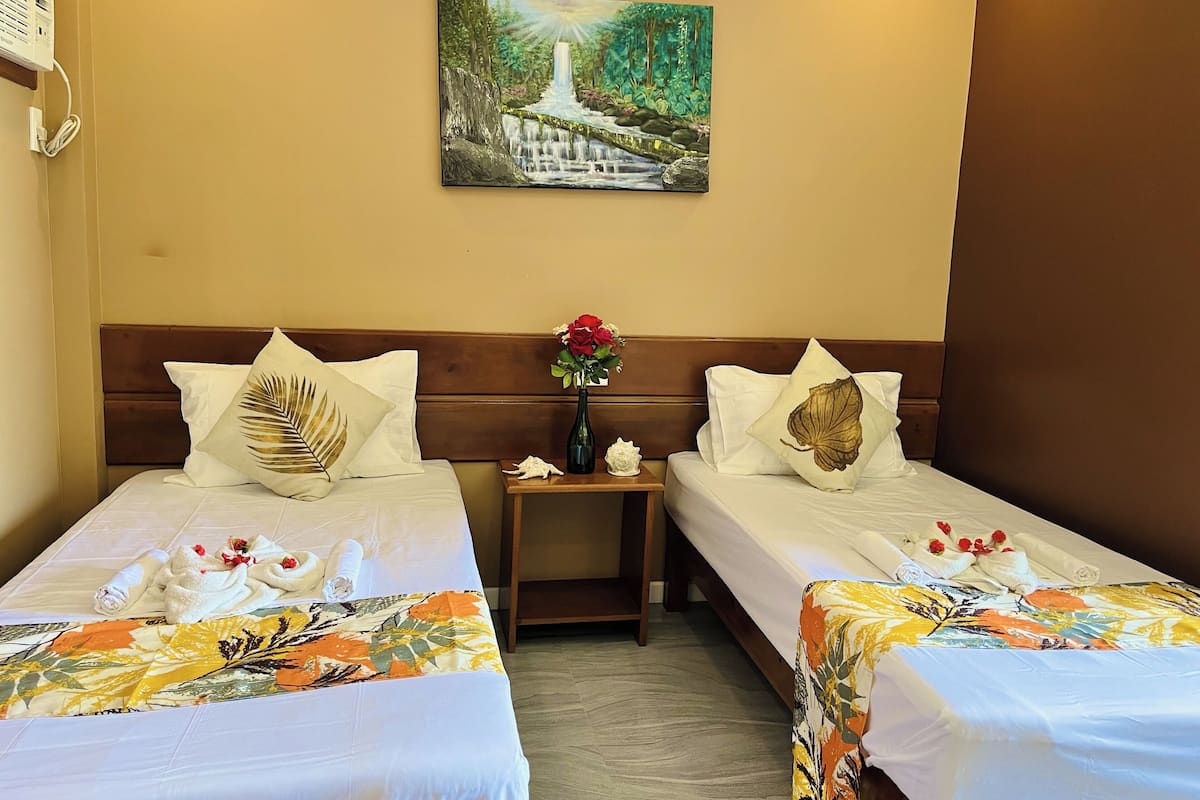 A hotel room with two single beds, a painting of a waterfall, a nightstand with a vase of flowers, and a wooden headboard.